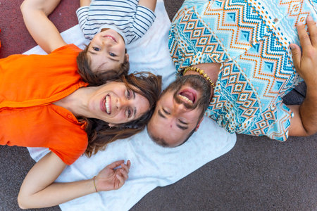 A family of three, a man, a mother and a son, are lying on a blanket on the ground. The man is wearing a blue and white shirt with a pattern of diamonds. The children are smilingの写真素材