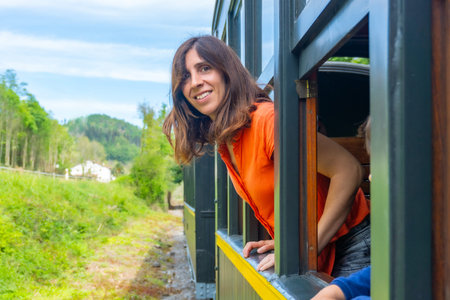 A woman is sitting in a train car with her head out the window. She is smiling and looking out at...