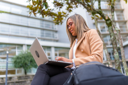 Side view low angle of a latin confident businesswoman using laptop sitting on city benchの写真素材