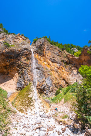 Majestic rinka waterfall flows down rocky cliffs in Logarska dolina valley, creating a picturesque scene amidst lush greenery under a vibrant blue skyの写真素材