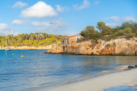 Tranquil turquoise waters lapping against the shore of s'amarador beach, with a weathered building atop a rocky outcrop surrounded by lush greenery under a serene blue skyの写真素材