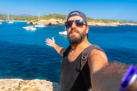 A man with a beard and sunglasses is taking a selfie in front of a body of water. The water is blue...