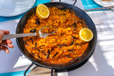 Close up of a spoon scooping up seafood paella from a traditional paella pan, garnished with lemon wedges, during a summer mealの写真素材