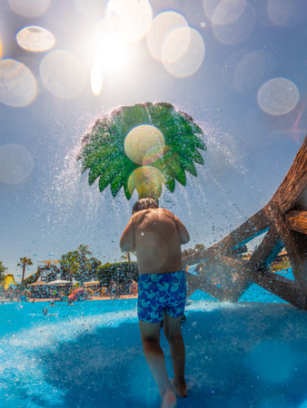 Child playing joyfully under a palm tree shaped shower at a vibrant water park during a sunny summer vacation in Alicante, Spainの写真素材