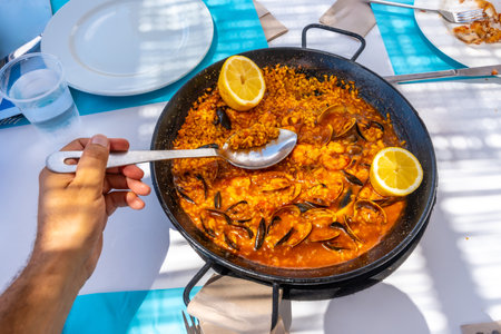 Hand holding spoon scooping seafood paella from pan in outdoor restaurant during summer vacationの写真素材