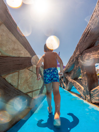 Young boy enjoying summer vacation, walking on blue water slide under bright sun at a water parkの写真素材