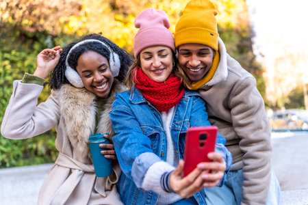 Three young multi ethnic friends wearing winter clothes are taking a selfie with a smartphone in an...