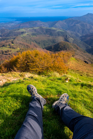 Hiker resting on grassy mountaintop, enjoying breathtaking views of rolling hills, colorful foliage, and distant ocean in autumnの写真素材
