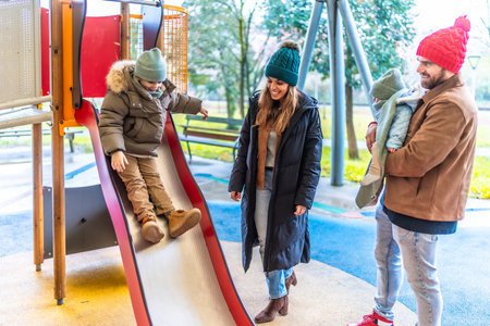 Cheerful family enjoying a winter day at the playground, with their son sliding down and parents lovingly watchingの写真素材