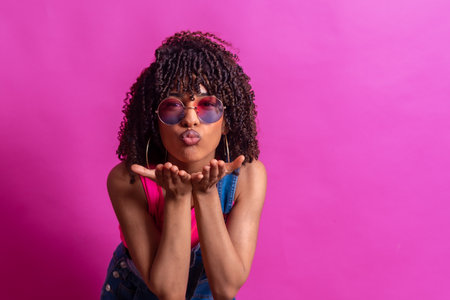 Fashionable young woman blowing a kiss while wearing sunglasses, set against a vibrant pink backdropの写真素材