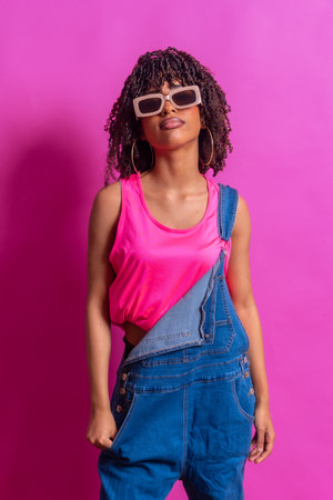 Fashionable woman with curly hair wearing trendy sunglasses, a vibrant pink top, and denim dungarees, confidently posing against a bright pink backdropの写真素材
