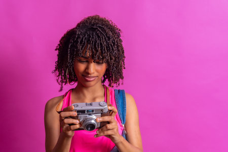 Young woman with curly hair and backpack looking at her vintage camera against a vibrant pink backgroundの写真素材