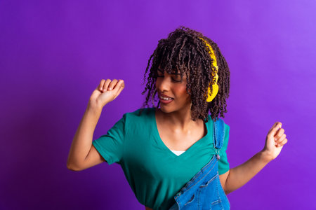 Happy young woman with yellow headphones dancing and enjoying music on a vibrant purple background, expressing joy and carefree attitudeの写真素材
