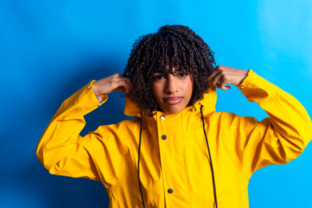 Studio shot of a young woman with curly hair adjusting the hood of her yellow raincoat against a vibrant blue backdropの写真素材