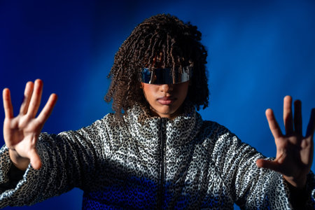 Young woman with futuristic visor and patterned jacket pushing screen on blue backgroundの写真素材