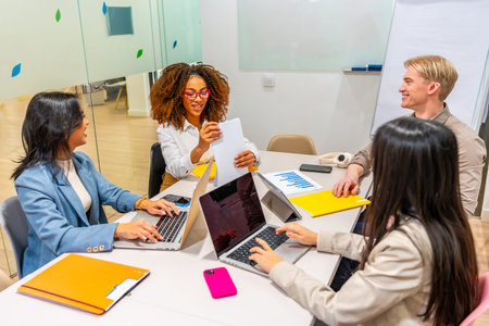 Four diverse colleagues are working together, using laptops and analyzing documents, showcasing...