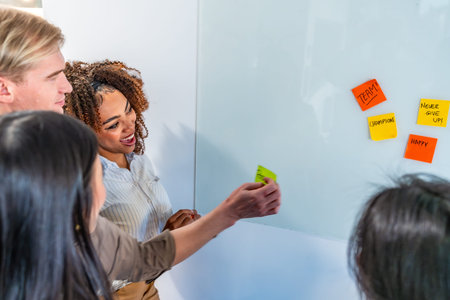 Office workers adding sticky notes with positive messages on whiteboard during a brainstorming...