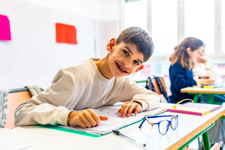 Smiling pre adolescent boy studying at his desk in elementary school classroom, showing inclusion and diversity in educationの写真素材