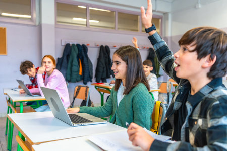 Elementary school students raising hands and using laptops in an interactive classroom, engaging in modern educational activitiesの写真素材