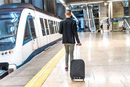 Businessman walking away with a trolley bag on the platform of a subway station, navigating the bustling urban environment during daytimeの写真素材