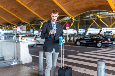 Young businessman using his smartphone while waiting for a taxi outside airport an terminal, holding a reusable coffee cup and pulling a suitcaseの写真素材