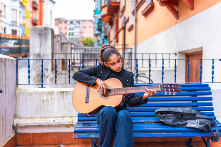 African american woman playing acoustic guitar sitting on a blue bench in the city