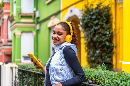 Happy african american woman using phone and listening to music in the city next to colorful housesの写真素材