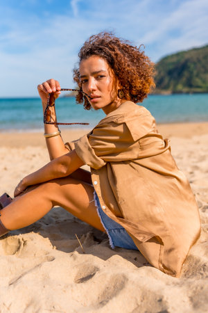 Vertical side view of a beauty Arab model sitting on sandy beach taking off the sunglasses and looking confident at cameraの写真素材