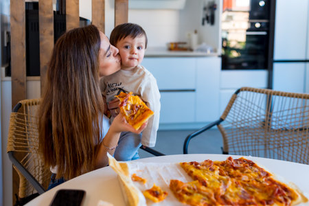 Mom kissing her son while enjoying a pizza lunch together in a modern kitchen, sharing moments of love and happiness in their homeの写真素材