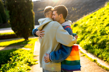 Two men embracing in a park, one holding a rainbow tote bag and a disposable cup, radiating love and happiness during golden hourの写真素材