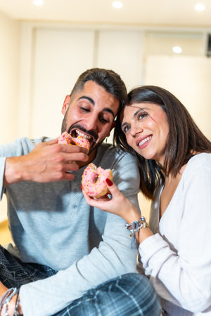 Young couple sitting on the floor at home, eating pink donuts with sprinkles, having fun and enjoying their time togetherの写真素材
