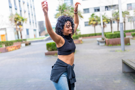 Curly haired woman dancing with arms raised in urban street, wearing black top and blue jeans