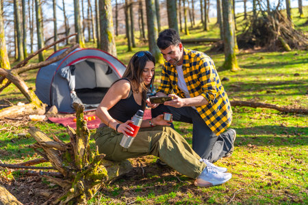 Couple of hikers enjoying a camping trip in the woods, watching photos on their smartphone while drinking hot beveragesの写真素材
