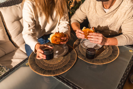 Couple sharing a relaxed moment, eating burgers and drinking beverages at a glass table, creating a cozy and intimate atmosphereの写真素材