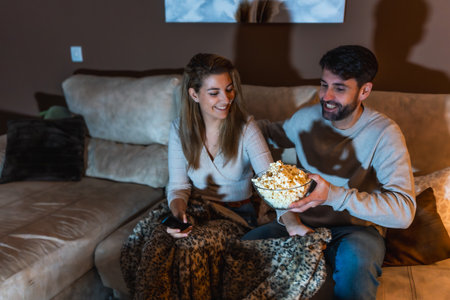 Happy couple sharing popcorn and enjoying a cozy movie night at home, snuggled together on the sofa under a warm blanketの写真素材