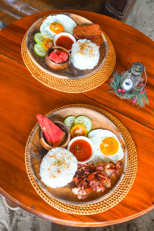 Two plates featuring Filipino breakfast with fried eggs, rice, meat, watermelon, tomato, cucumber, and hot sauce on a wooden table in Siargao island, Philippinesの写真素材