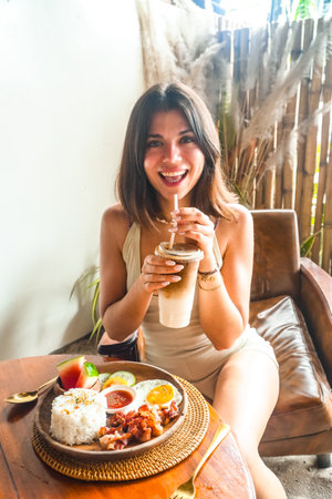 Happy tourist sipping iced coffee while enjoying a traditional Filipino breakfast of bacon, eggs, rice, and fresh fruit in Siargaoの写真素材