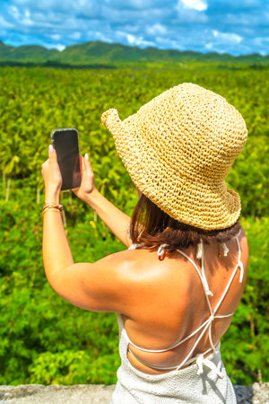 Young woman wearing a straw hat taking pictures of a coconut plantation in Siargao island, Philippines, during a sunny summer dayの写真素材