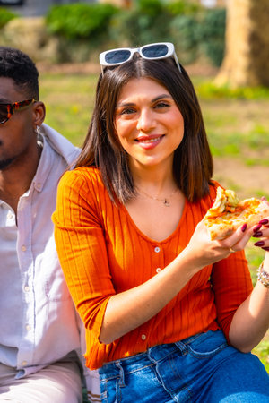 Smiling young woman enjoying a slice of pizza outdoors in a park, sitting next to a friend, wearing...