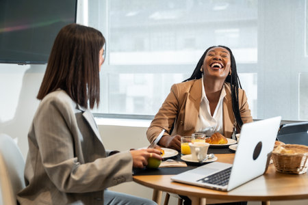 Two businesswomen are enjoying breakfast and laughing together during their business trip, creating a positive and productive atmosphereの写真素材