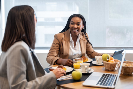 Two businesswomen enjoying breakfast and engaging in conversation during their business trip, using laptop and tabletの写真素材