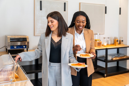 Two businesswomen are smiling while having breakfast at a hotel buffet, showing a positive and productive start to their business tripの写真素材