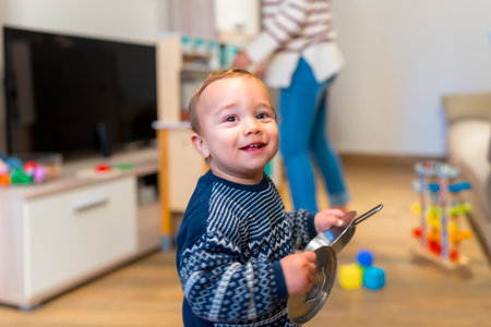 Smiling toddler playing with kitchenware in the living room while mother is tidying up in the background, concept of happy family lifeの写真素材