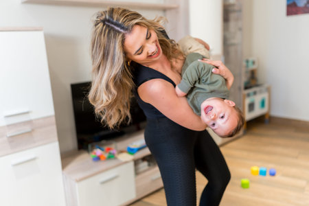 Cheerful mother lifting and playing with her baby boy, having fun together in the living room