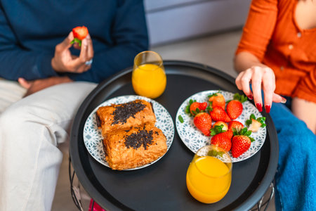 Multiethnic couple sharing a delightful breakfast at home, savoring pastries, fresh strawberries, and refreshing orange juiceの写真素材