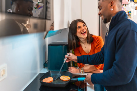 Cheerful multiethnic couple laughing and having fun while preparing burgers together on the stovetop in their modern kitchenの写真素材