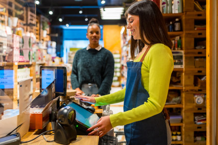 Smiling saleswoman wearing apron using touchscreen cash register while customer waiting at checkout...
