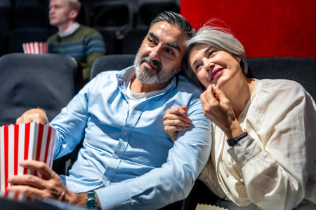 Senior couple enjoying a cozy movie night at the cinema, savoring popcorn while sharing a heartfelt moment filled with joy and affectionの写真素材