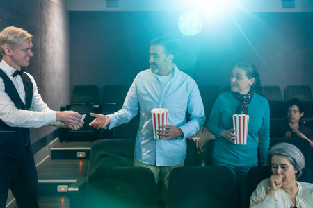 Senior couple holding popcorn buckets entering movie theater and receiving tickets from usher,...