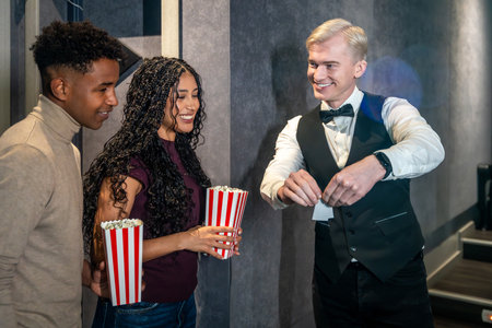 Young couple holding popcorn buckets entering a movie theater and checking their tickets with the usher before watching a filmの写真素材